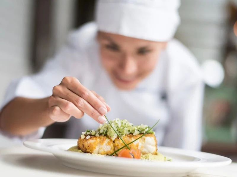 A chef in a white uniform and hat is garnishing a plated dish with herbs, carefully placing them on top of a piece of fish, focusing intently on the final presentation.