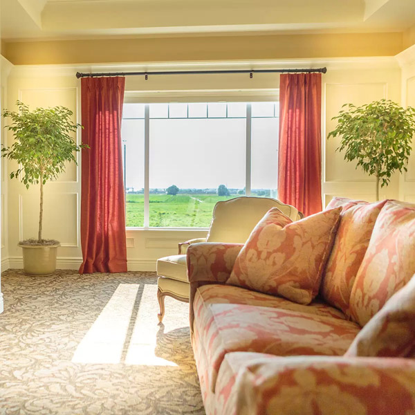 A cozy living room with a floral-patterned sofa and armchair, large window with red curtains, two potted plants, and sunlight streaming in, overlooking a green field outside.