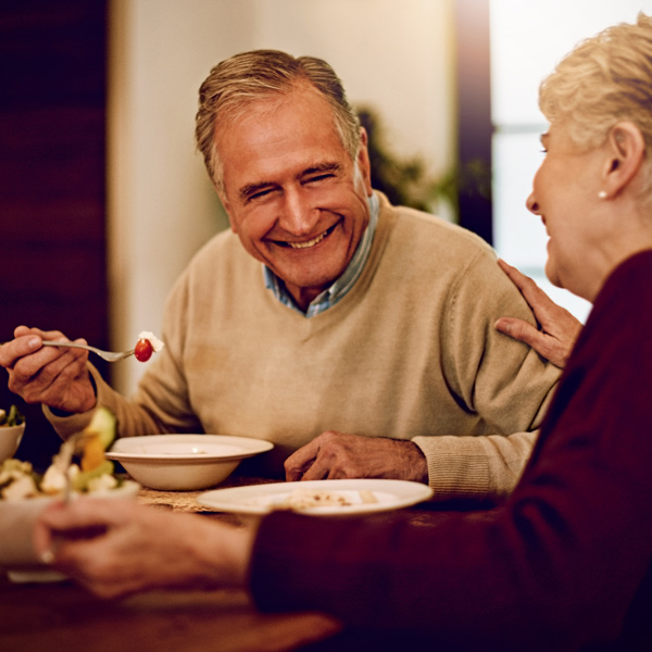 An older man and woman sit at a dining table, smiling and enjoying a meal together. The man holds a spoon with food, while the woman touches his shoulder, both appearing happy and engaged in conversation.