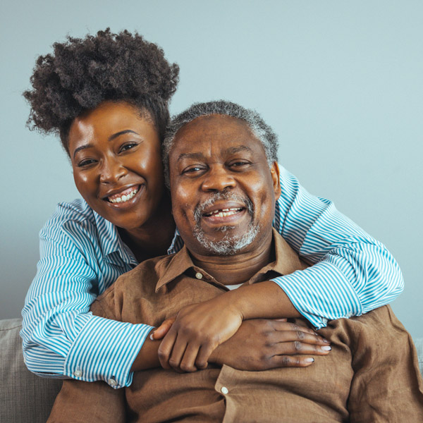 A smiling young woman hugs an older man from behind as they sit on a couch, both looking at the camera. The background is a plain light blue wall.