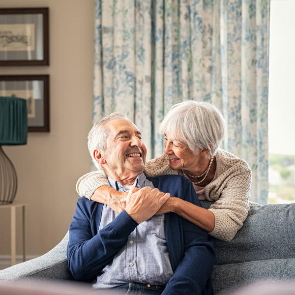 An elderly couple sits on a couch at home, smiling and embracing warmly. The woman hugs the man from behind, and both look happy and relaxed in a cozy, well-lit living room.