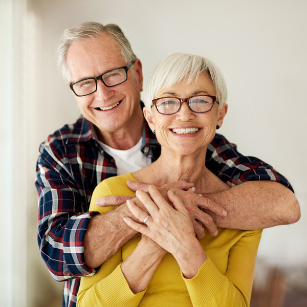 An older couple, both wearing glasses and smiling, stands together indoors. The man hugs the woman from behind, and she clasp his arms, both appearing happy and relaxed.