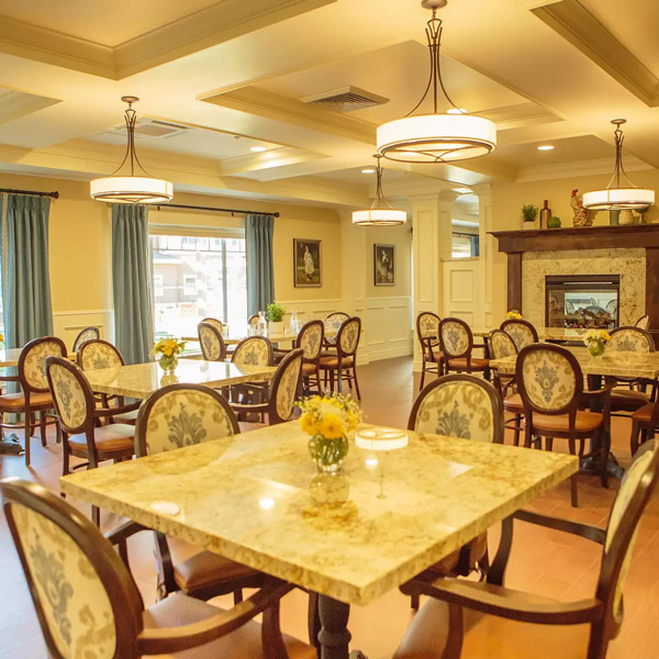 A bright, elegant dining room with marble tables, floral-upholstered chairs, fresh flower centerpieces, large windows with blue curtains, paintings on the walls, and a fireplace with decorative items above it.