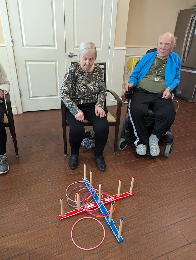 Two elderly adults sit indoors, focusing on a ring toss game on the floor with colorful rings and pegs. One woman is seated on a chair, the man is in a wheelchair, both participating in the activity.