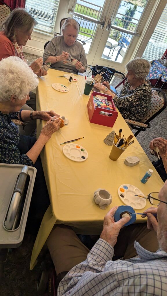 Five older adults sit around a yellow-covered table, painting pottery pieces with brushes and paint palettes. Art supplies and a red tissue box are on the table. Sunlight shines through windows in the background.