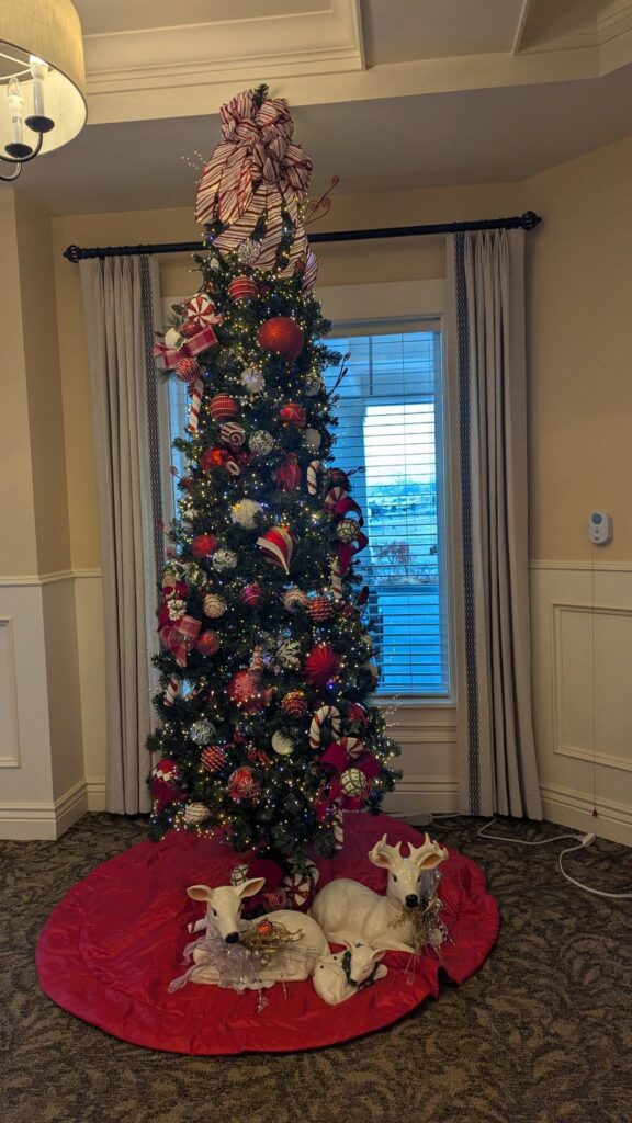 A decorated Christmas tree with red and white ornaments stands in front of a window, topped with a large striped bow. Two white decorative reindeer rest on a red tree skirt at the base.