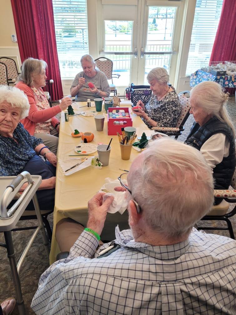 Six elderly people sit around a table engaging in arts and crafts activities, with paints, brushes, and supplies spread out. They appear focused and are seated in a bright room with large windows.