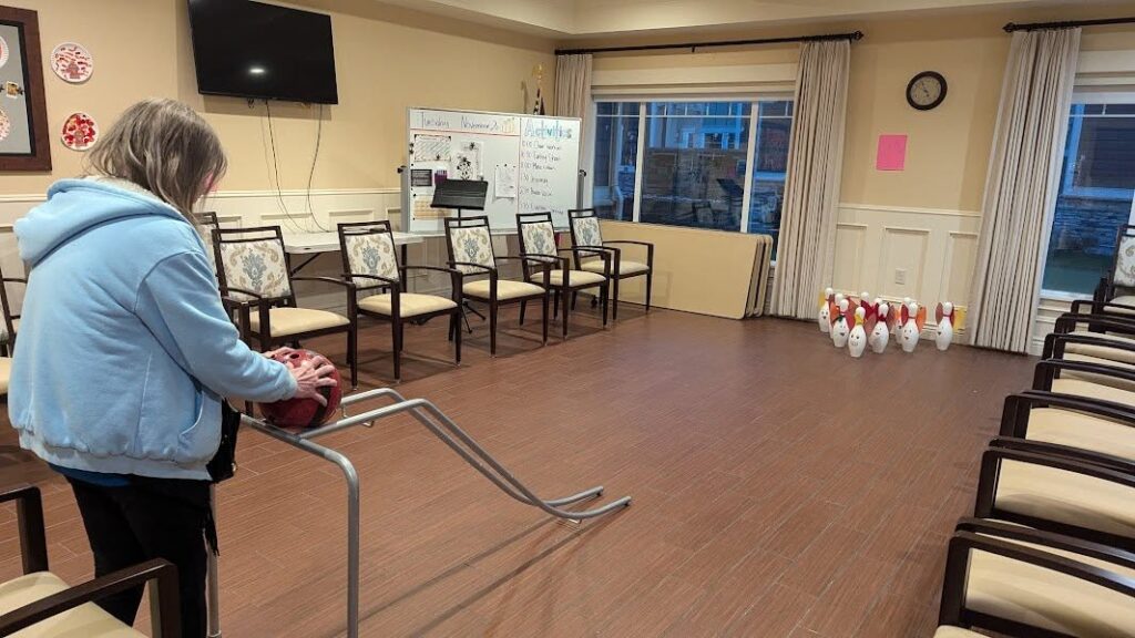 A person stands next to a metal bowling ramp, aiming a bowling ball toward ten plastic pins set up at the far end of a room with chairs arranged along the walls.