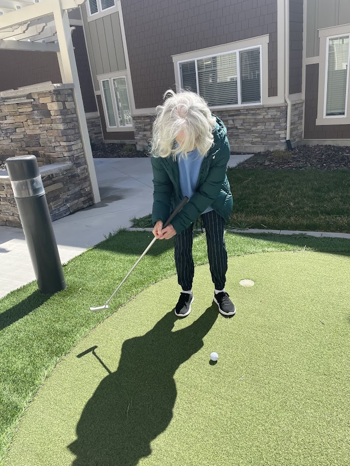 An older adult with gray hair, wearing a green jacket and striped pants, prepares to putt a golf ball on a small artificial green outside a residential building on a sunny day.