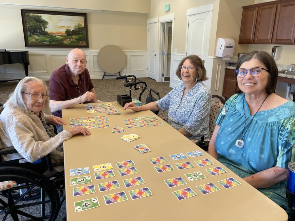 Four elderly adults sit around a table, smiling and playing a colorful card game in a bright, communal room with a painting on the wall and a kitchen area in the background.