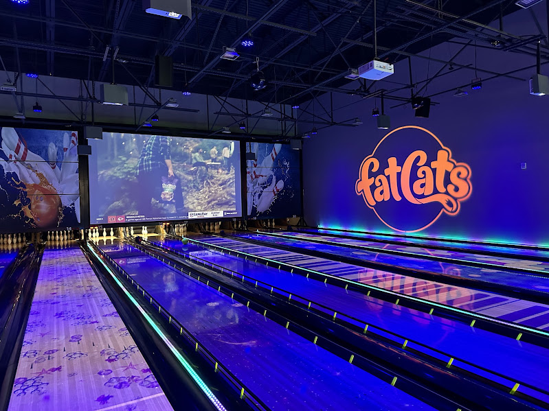 A neon-lit bowling alley with several lanes, a large "FatCats" logo on the wall, and a projection screen displaying an image at the end of the lanes.