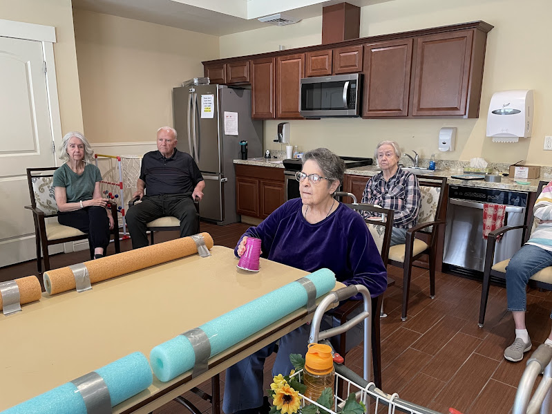 A group of elderly people sit spaced apart in a kitchen area, some with walkers and pool noodles on the table, while one woman in the foreground holds a pink can. The setting appears to be a care facility or senior home.