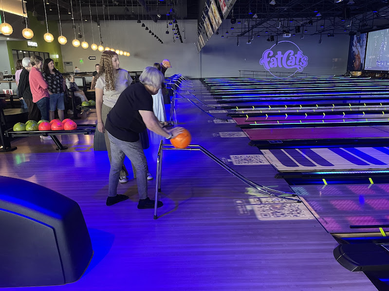 An older woman uses a metal ramp to roll a bowling ball down a lane at a bowling alley, with several people watching nearby. The lanes are lit with colorful neon lights, and “Fat Cats” is displayed on the back wall.