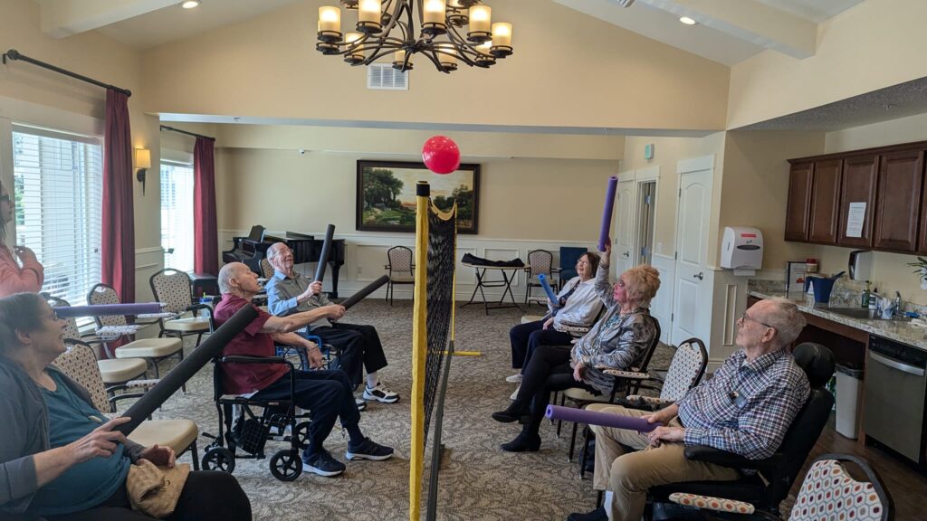 A group of older adults sits in a circle in a bright room, using foam noodles to hit a red balloon suspended in the air, engaging in a seated group activity.