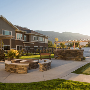 A landscaped backyard features a stone fire pit with circular seating, a wooden bench, and a pergola, with a large house and mountains in the background under a clear sky.