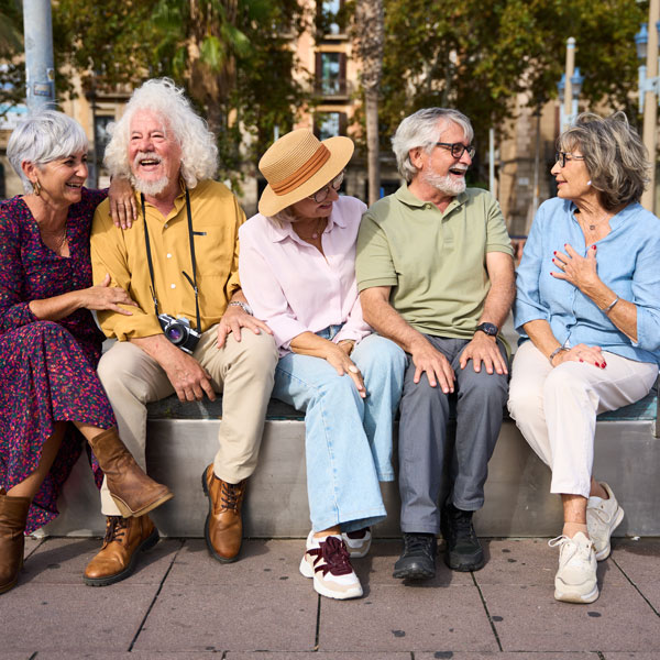 Five older adults sit together outdoors on a bench, laughing and chatting. They wear casual clothing and look happy, with trees and sunlight in the background.