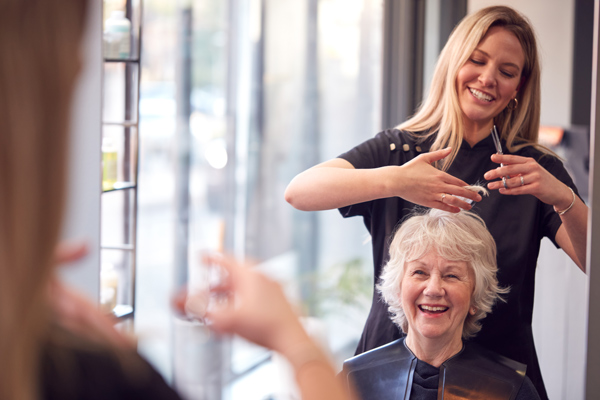 A smiling older woman sits in a salon chair with a black cape as a hairstylist stands behind her, styling her hair. Both women appear happy and are looking in the mirror.