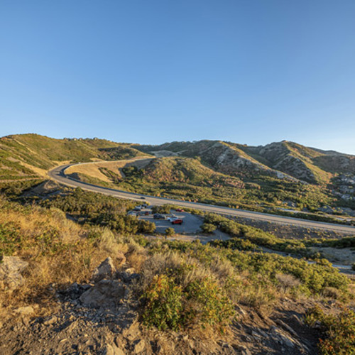 A winding road cuts through green, rolling hills under a clear blue sky. A small parking lot with several cars is visible near the road, surrounded by lush vegetation and rugged terrain.