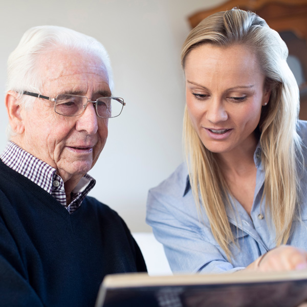An elderly man wearing glasses sits next to a younger woman with long blond hair. They are both looking at a photo album together, appearing engaged and interested.