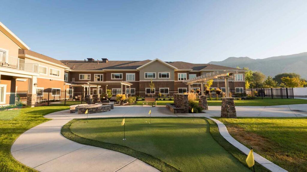 A well-maintained courtyard features a small putting green with yellow flags, surrounded by walkways, stone planters, and a large two-story building in the background, with mountains visible under a clear sky.