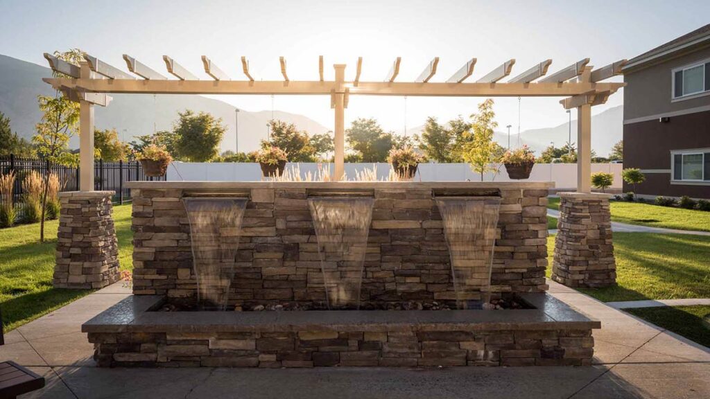 Stone fountain with three waterfalls in a landscaped yard, backed by a white pergola with potted plants on top. Sunlight streams through the pergola, and a house and mountains are visible in the background.