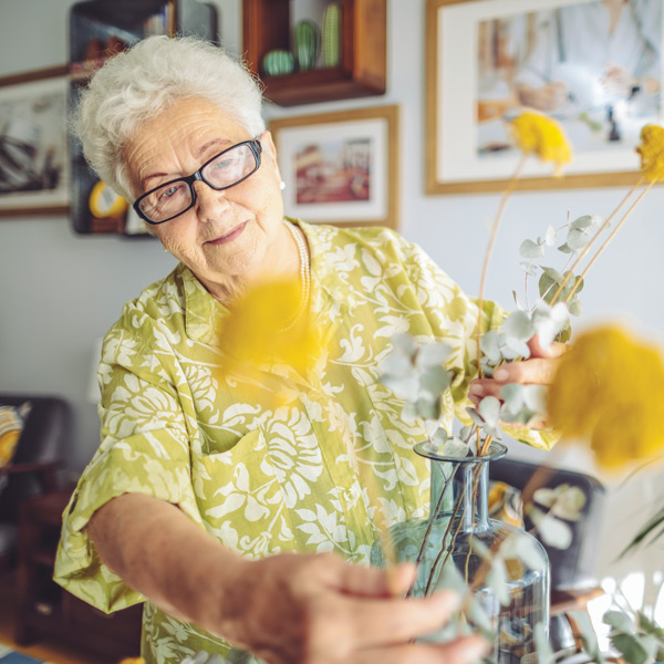 An elderly woman with short white hair and glasses arranges yellow flowers in a vase. She is smiling and wearing a green floral shirt. The background shows framed pictures and home decor.