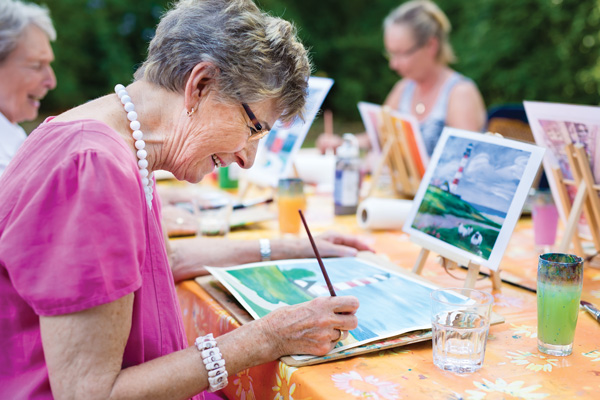 An elderly woman wearing glasses and a pink shirt paints on a canvas at an outdoor table, smiling. Other people are also painting in the background. The table is covered with art supplies and drinks.