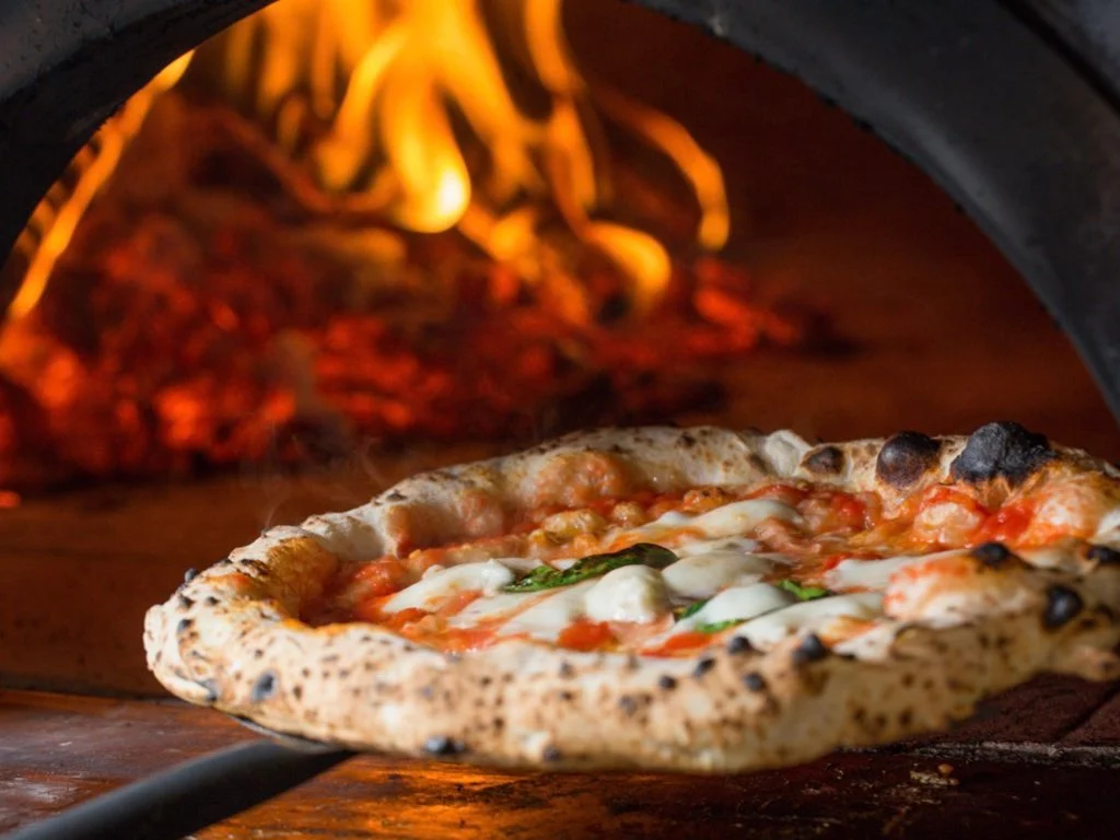 A freshly baked pizza with melted cheese and basil is being pulled from a wood-fired oven, with flames and glowing embers visible in the background.