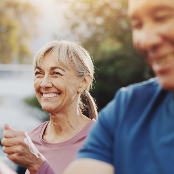 Smiling older woman in a pink top outdoors, with another person in a blue shirt out of focus in the foreground, both appearing happy and active in natural sunlight.