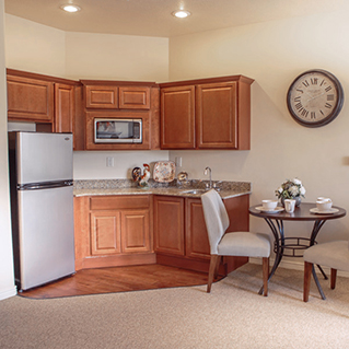 A small kitchen with wooden cabinets, a stainless steel fridge, microwave, granite countertops, and a round dining table with two upholstered chairs. A large clock hangs on the wall above the table.