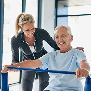 A smiling older man in a light blue shirt exercises with a blue resistance band, assisted by a woman in sportswear who is also smiling. They are in a bright, modern room with large windows.