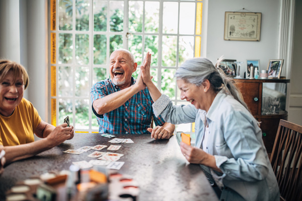 Three older adults sit around a table playing cards, laughing and giving a high five. Sunlight streams through large windows behind them, and the room appears cozy and inviting.