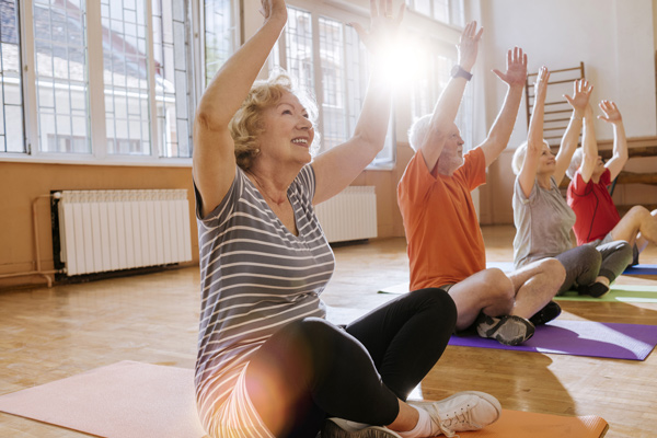 Four older adults sit on yoga mats in a bright, sunlit room, raising their arms overhead and smiling during a group exercise or yoga class.