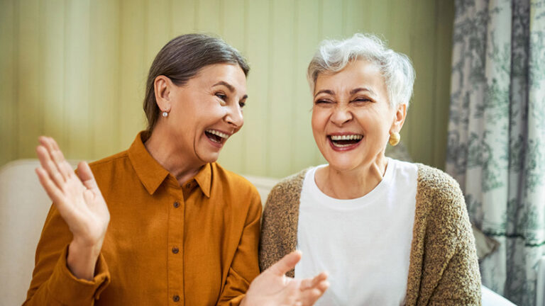 Two older women sit together indoors, smiling and laughing joyfully. Embracing the independent living philosophy, they enjoy each other's company—one in a mustard shirt gesturing as light streams in from a window behind them.