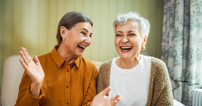 Two older women with gray hair are sitting indoors, smiling and laughing together. One wears an orange shirt and gestures with her hand, while the other wears a white shirt and cardigan. They appear joyful and happy.