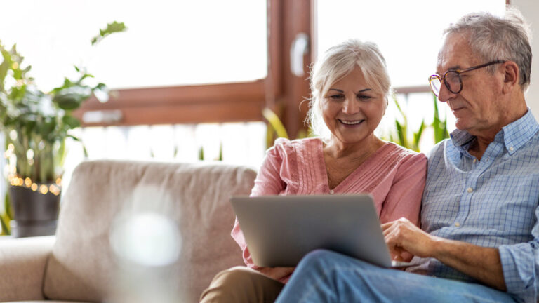 An older couple sits on a sofa at home, smiling while looking at a laptop together. Sunlight streams through the window, with green plants in the background—capturing the spirit of independent living philosophy.