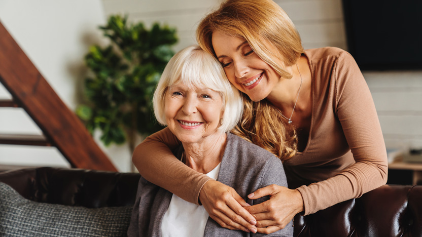 A young woman hugs an older woman from behind on a couch, both smiling and appearing happy. In the background, a plant and staircase add to the warm atmosphere. Their comfort and joy together highlight the positive impact of senior care technology in supporting family connections.